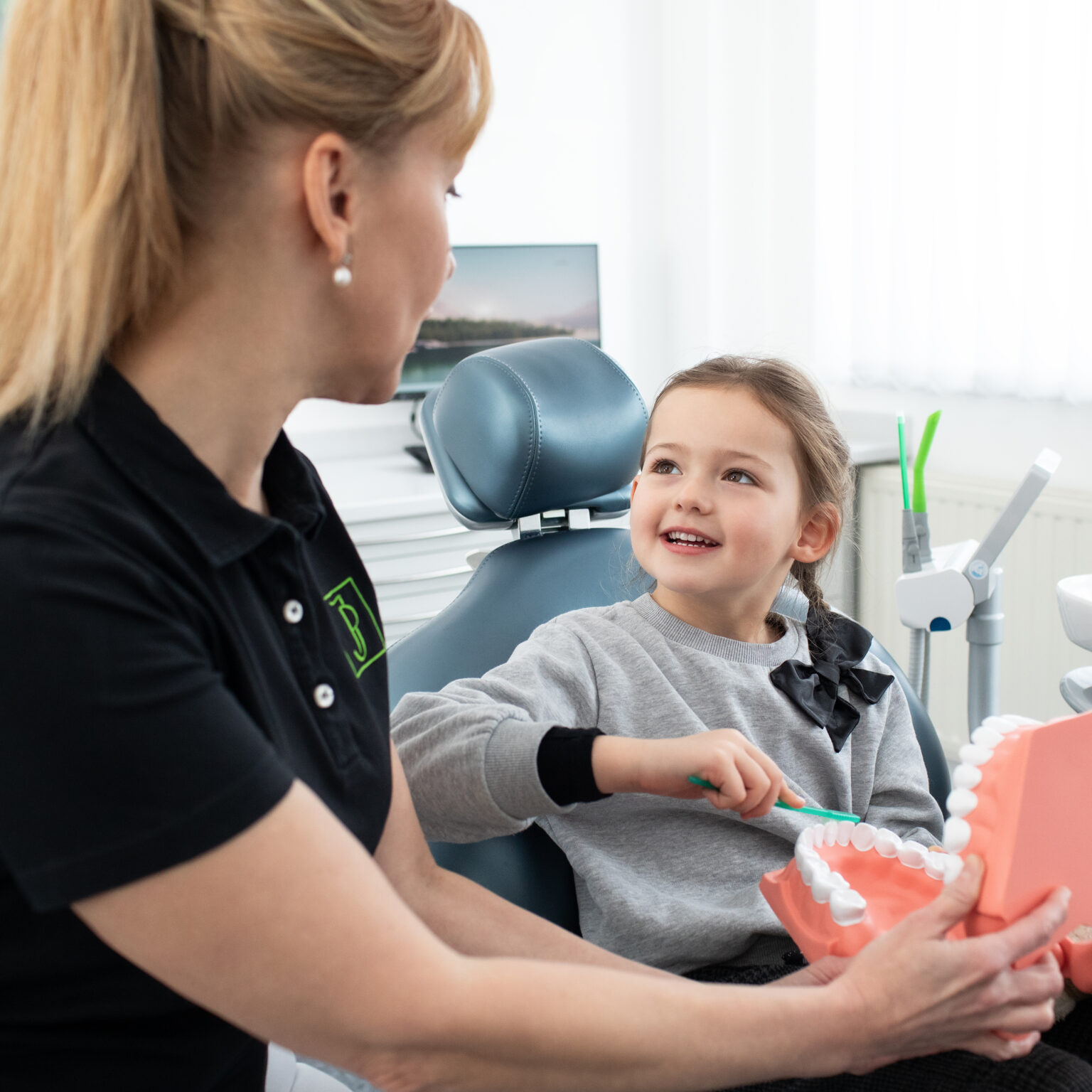 Zahnärztin erklärt einem lächelnden Mädchen anhand eines Zahnmodells die Kinderzahnheilkunde in der Zahnarztpraxis Pracht in Allershausen
